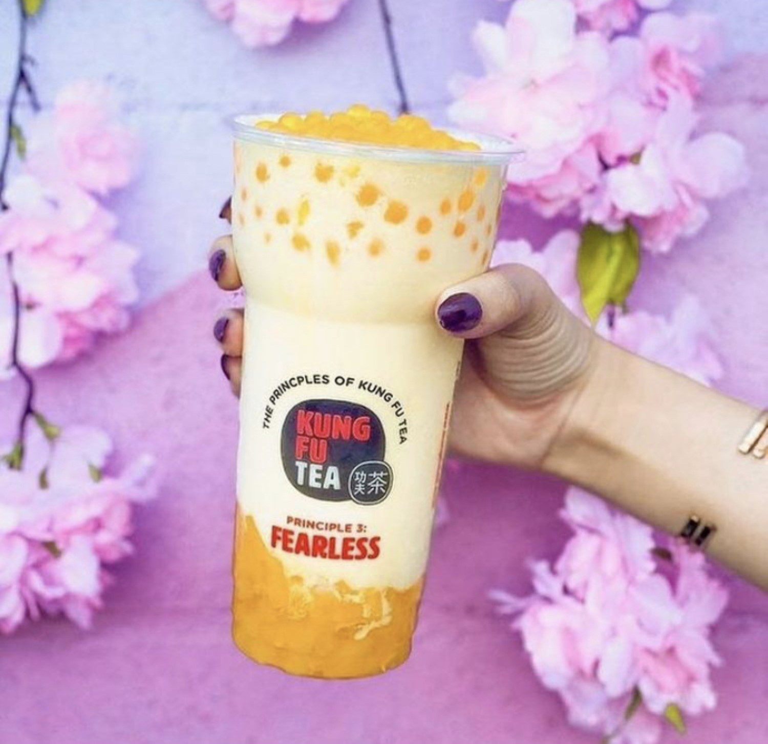 A hand holding a mango Kung Fu Tea drink against a cherry blossom background